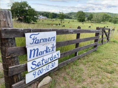 a white and blue sign on a wooden fence with a farm, a house, and cows in the background.