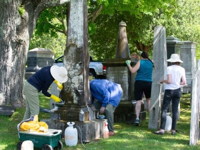 Two volunteers clean a tall gravestone with brushes while two more volunteers work on a momument behind them.