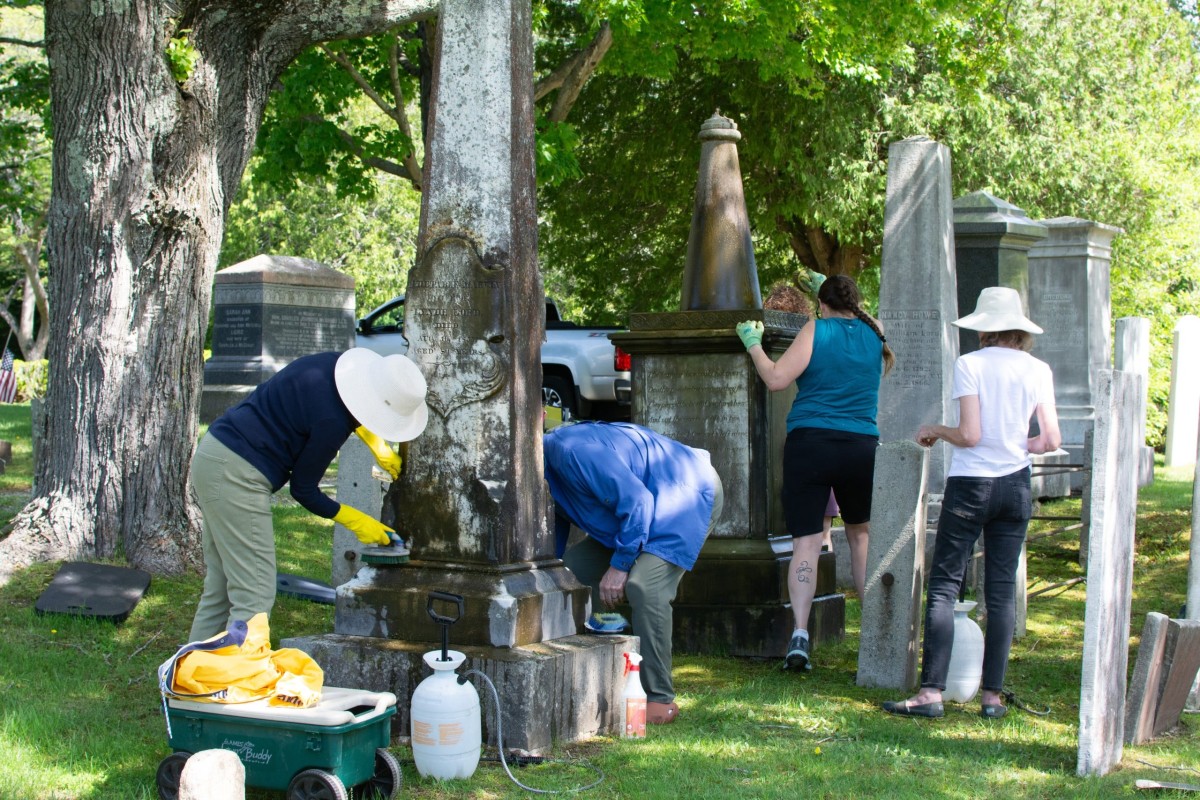 Two volunteers clean a tall gravestone with brushes while two more volunteers work on a momument behind them.