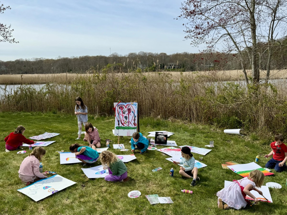 Kids sit on the grass while painting on posterboard along the river.
