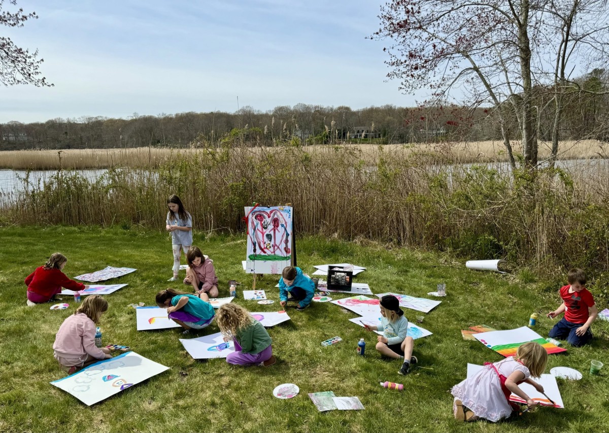 Kids sit on the grass while painting on posterboard along the river.