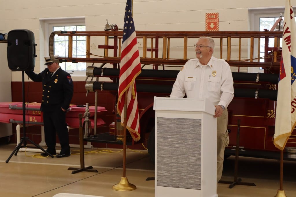 Roberge stands at the podium flanked by flags behind an antique fire truck.