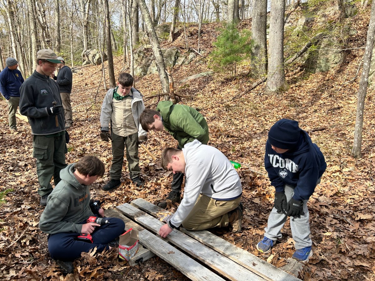 A Boy Scout bends over a section of the footbridge while five Boy Scouts look on.