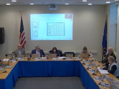 The board members are seated at tables set up in a U shape with a PowerPoint presentation projected behind them.