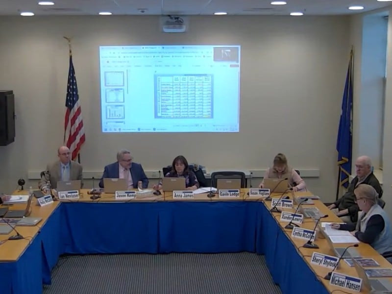 The board members are seated at tables set up in a U shape with a PowerPoint presentation projected behind them.