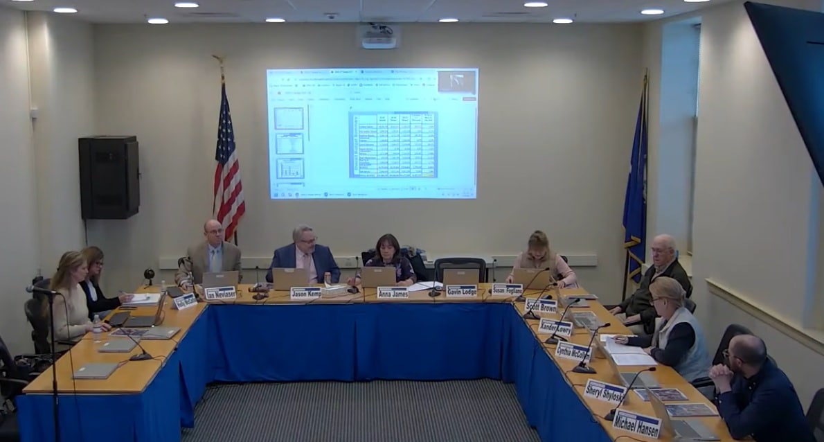 The board members are seated at tables set up in a U shape with a PowerPoint presentation projected behind them.