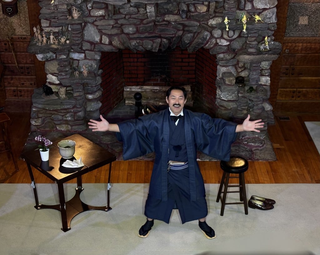 Hirai stands with arms outstretched in front of a fireplace.
