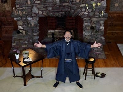 Hirai stands with arms outstretched in front of a fireplace.