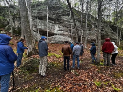 Hikers stand with their backs to the camera in front of a large rock outcropping and tall trees.