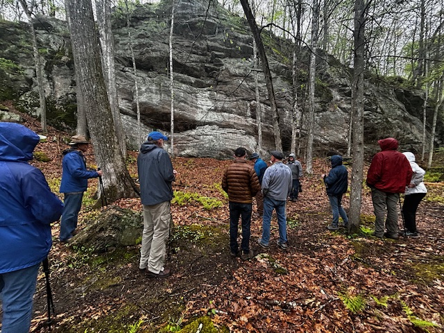 Hikers stand with their backs to the camera in front of a large rock outcropping and tall trees. 