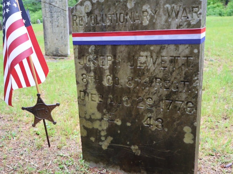 An hisotric gravestone next to a flag affixed to a star emblazoned with "1776"
