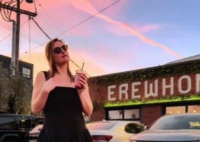 Artist holds an iced coffee while standing in front of a brick building with cars parked in front of it.