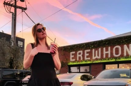 Artist holds an iced coffee while standing in front of a brick building with cars parked in front of it.