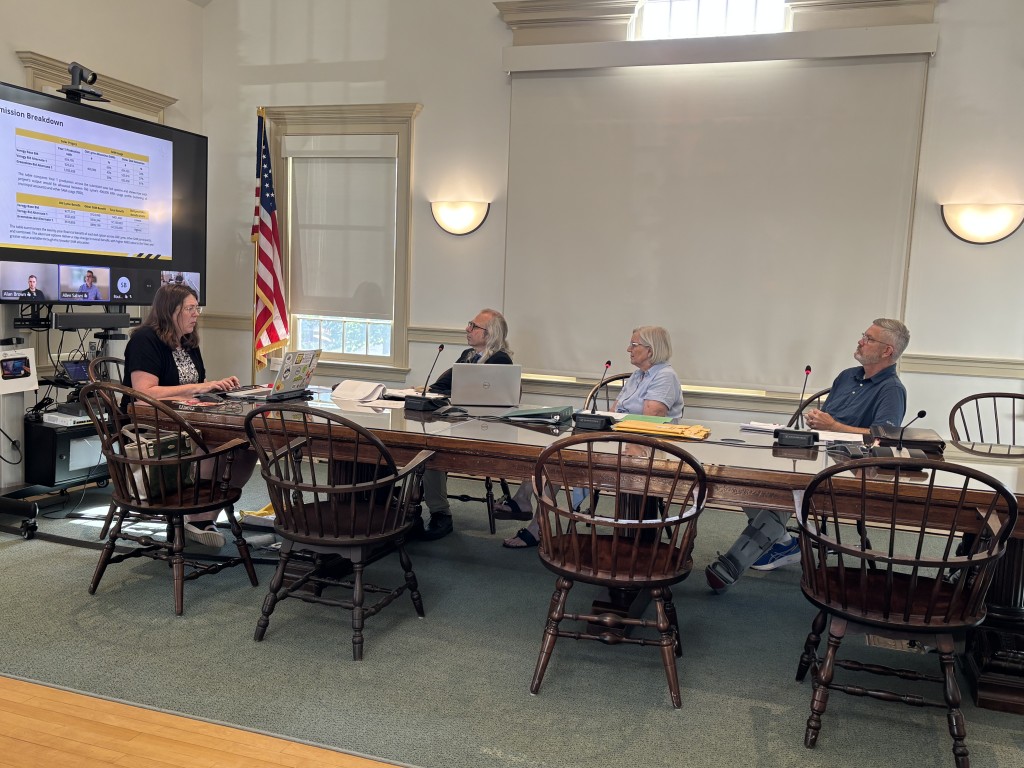 Three selectmen look at a computer screen showing the virtual presenters while the secretary takes notes.