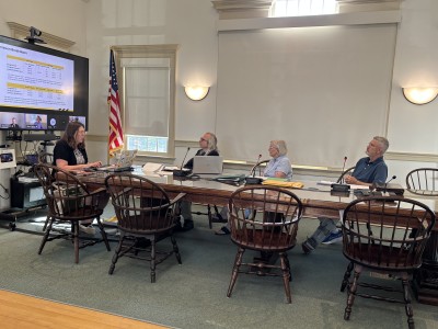 Three selectmen look at a computer screen showing the virtual presenters while the secretary takes notes.