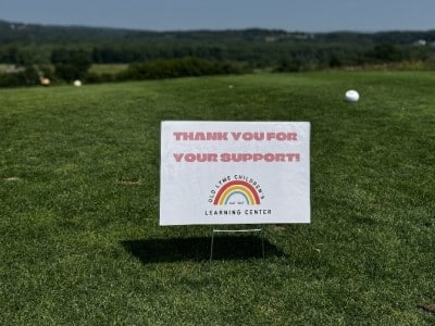 OLCLC sign thanking participants for their support sits on a golf course with a golf ball on the ground.