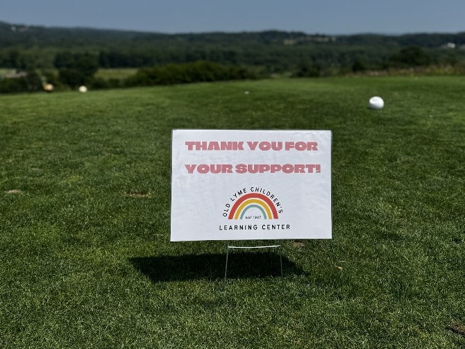 OLCLC sign thanking participants for their support sits on a golf course with a golf ball on the ground.