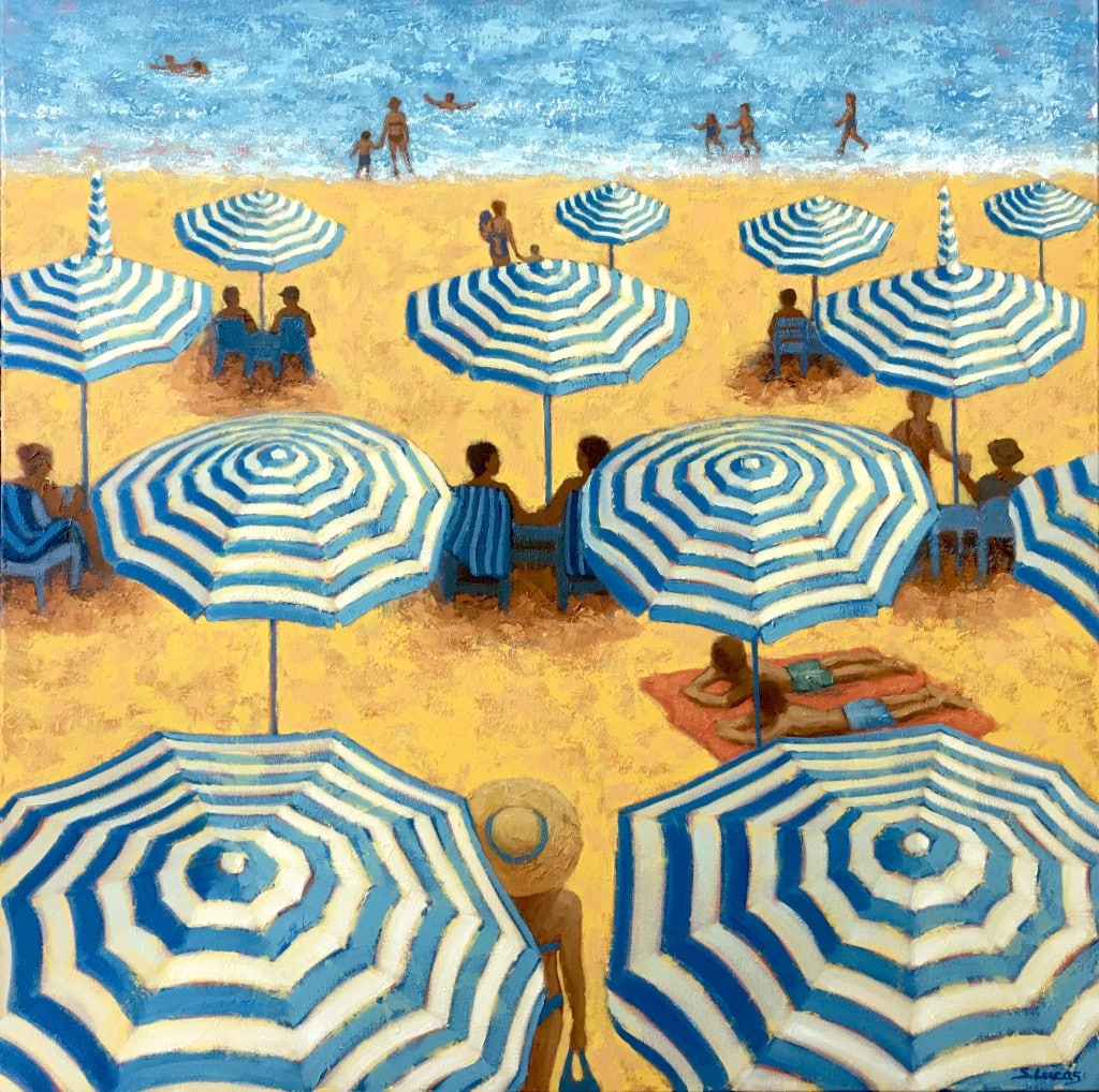 People sit under and around blue and white beach umbrellas on yellow-hued sand in front of blue water.