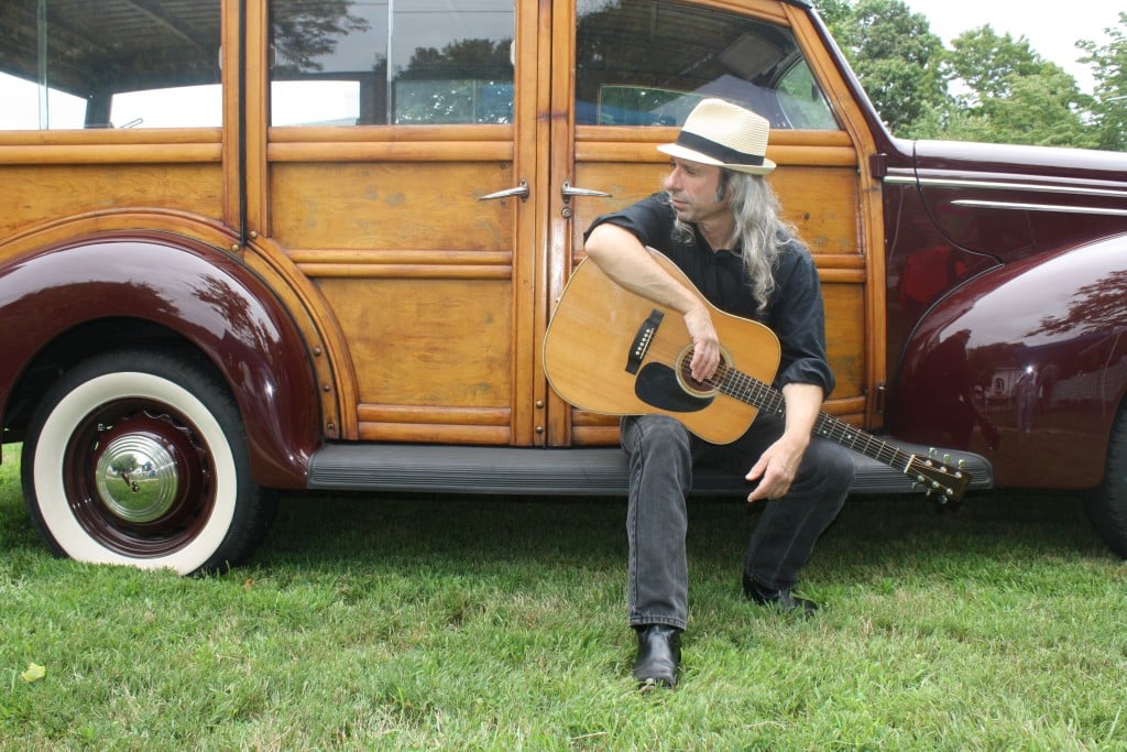 Artist with guitar sits on antique car running board. 