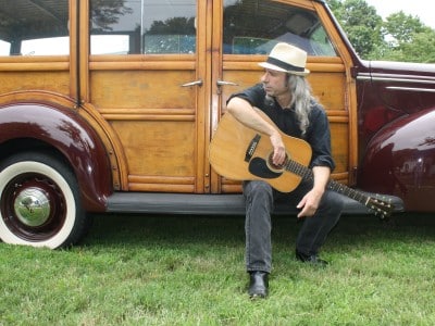 Artist with guitar sits on antique car running board.